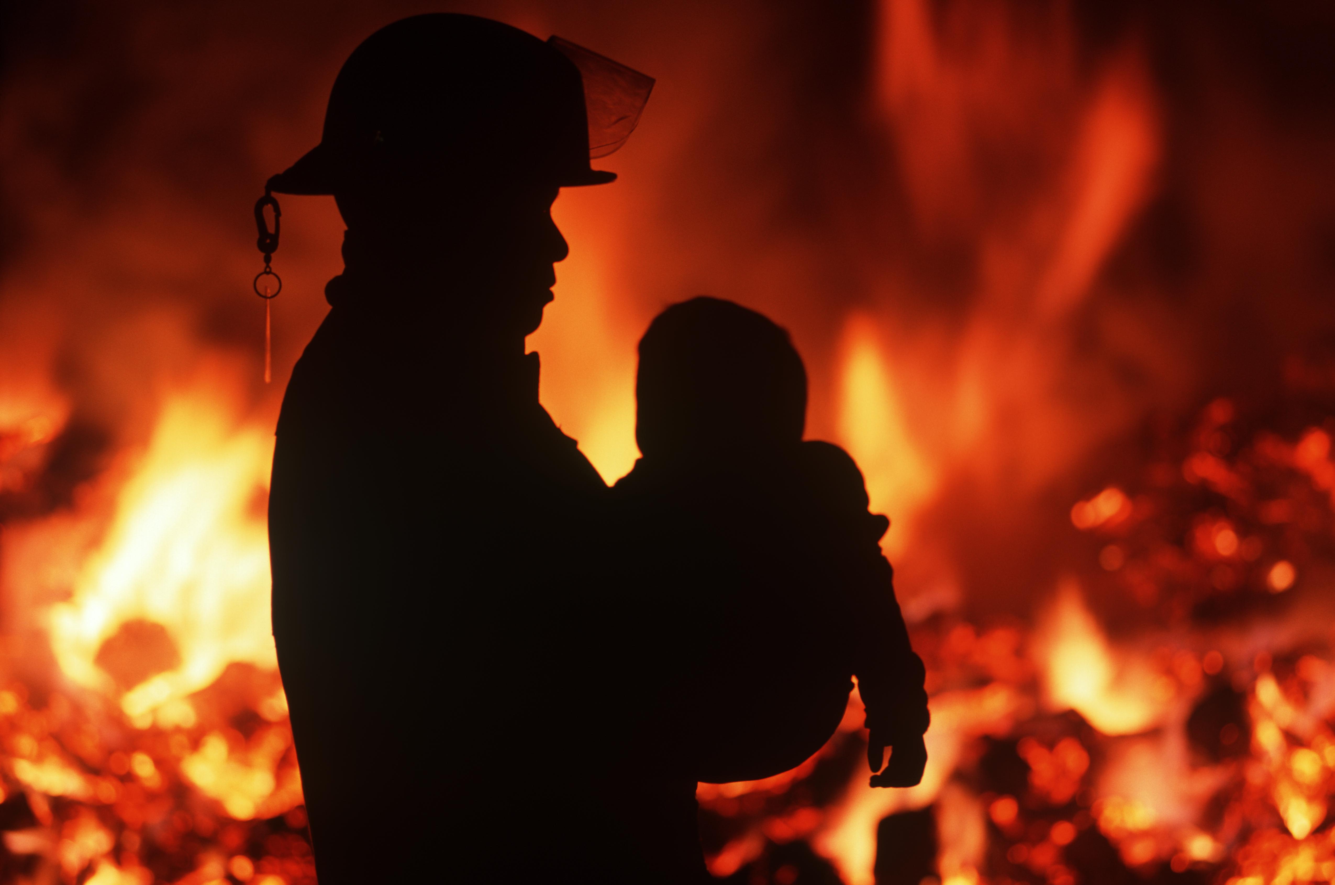 Silhouette of fireman carrying child with burning house beyond, British Columbia, Canada.