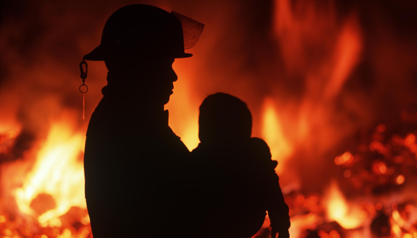 Silhouette of fireman carrying child with burning house beyond, British Columbia, Canada.