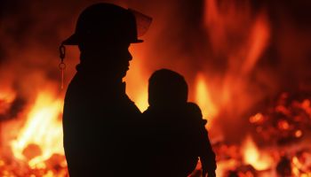 Silhouette of fireman carrying child with burning house beyond, British Columbia, Canada.