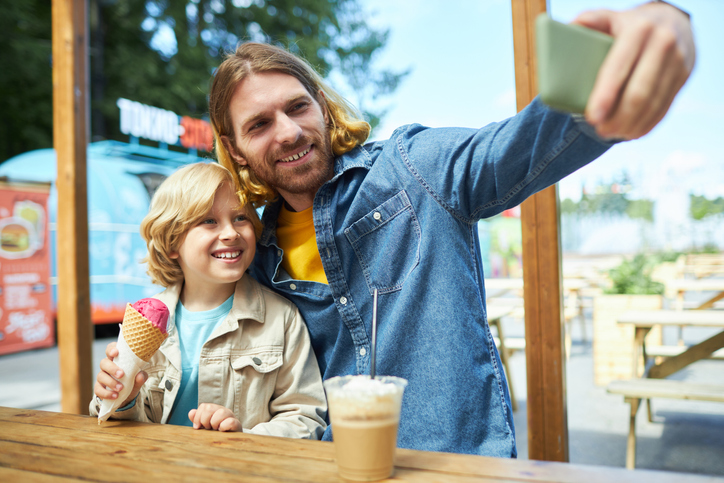 Father and son in amusement park eating ice cream and taking selfie