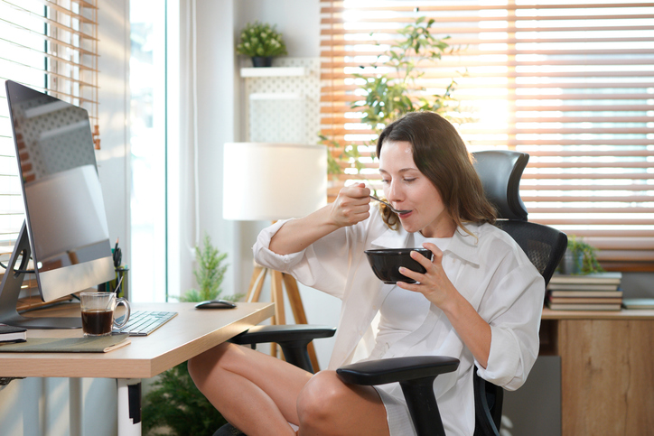 woman sitting at home office table has breakfast with desktop pc. Smiling relaxed young lady hybrid working on computer in modern house