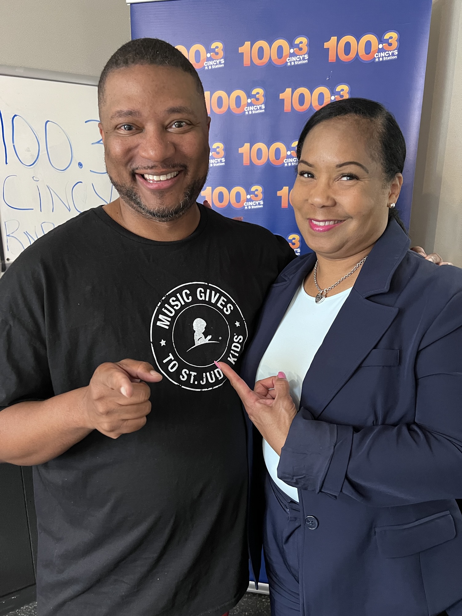 Two smiling people posing in front of a radio station backdrop with the call letters "100.3" visible.