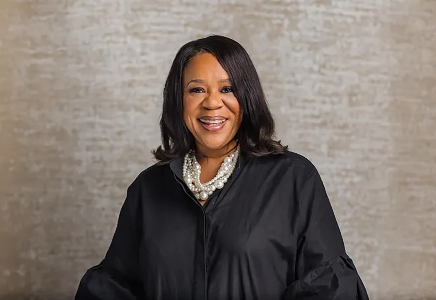 A smiling woman wearing a black jacket and a pearl necklace stands against a gray background.