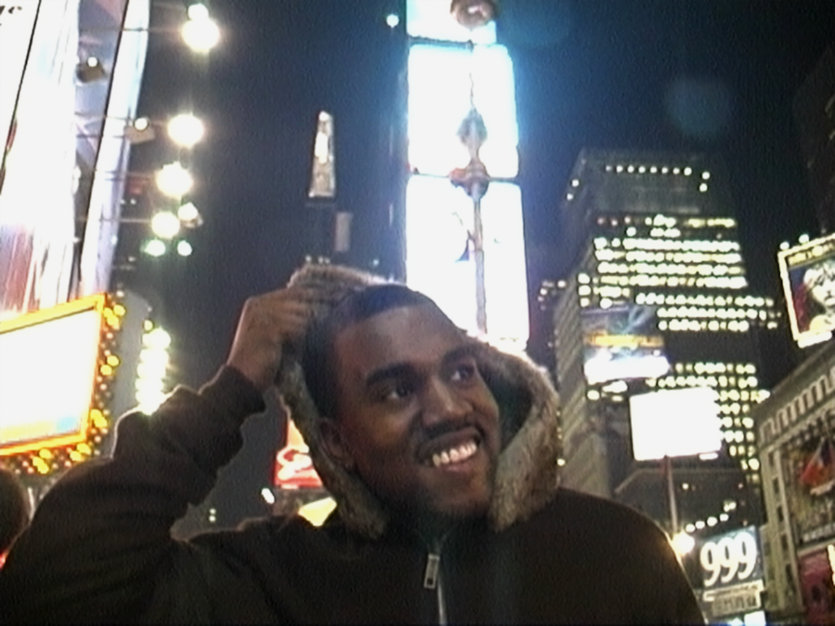 A smiling man with long hair and a beard stands in a brightly lit urban setting at night, with illuminated billboards and buildings visible in the background.