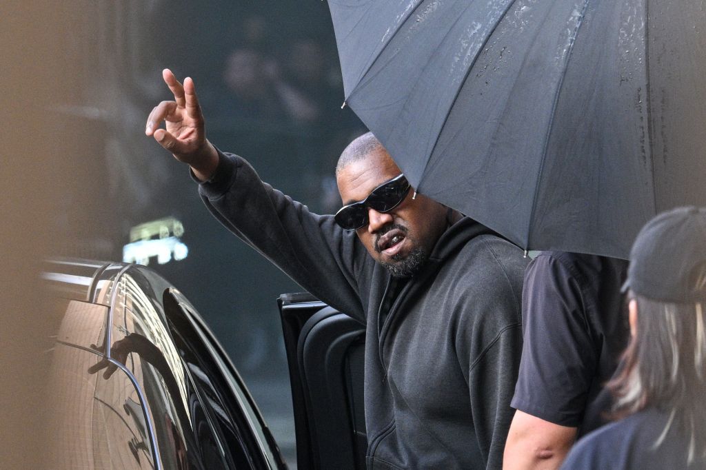 A person wearing sunglasses and a black hoodie gesturing a peace sign while holding an umbrella in front of a car.