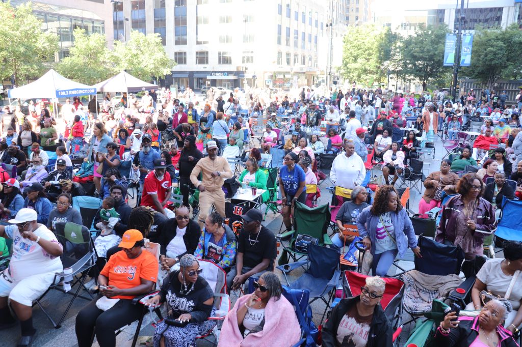 Crowded outdoor event with people sitting in chairs and tents, some wearing colorful clothing and accessories.