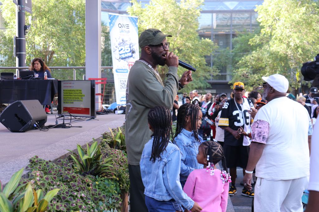 A man with a beard and hat speaking into a microphone while surrounded by a crowd, including children, in an outdoor setting with trees and buildings in the background.