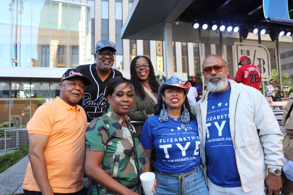 A group of six smiling people posing together outdoors, some wearing sports jerseys and hats with team logos.