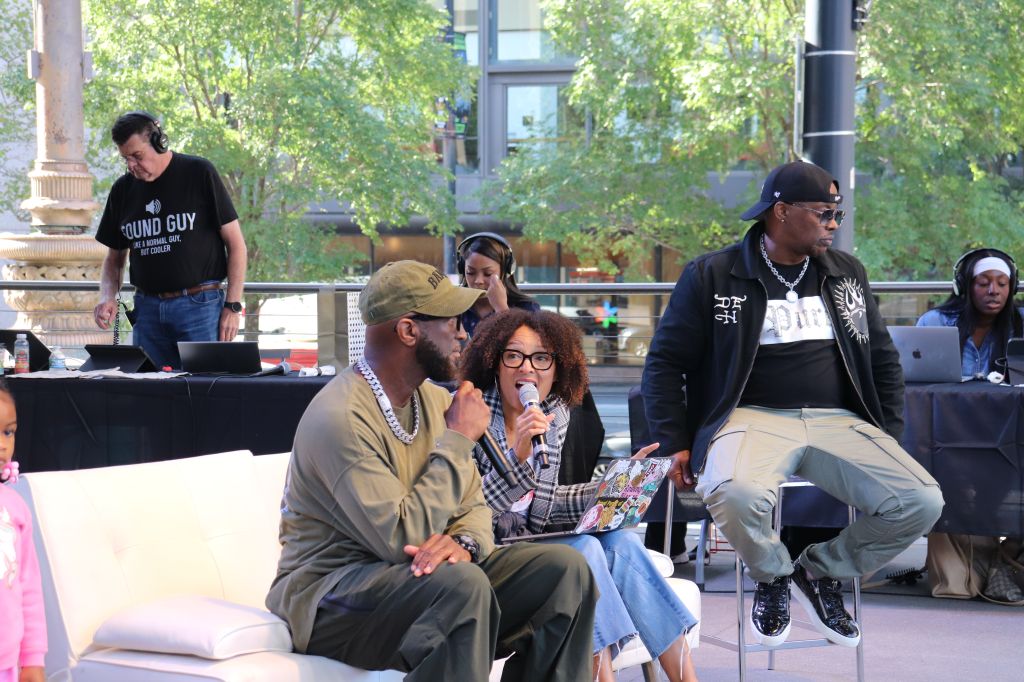 A group of people, including a woman holding a microphone, sitting on a stage in an outdoor setting with trees and buildings in the background.