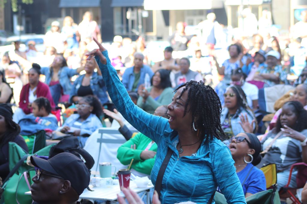 A crowd of people gathered outdoors, with a woman in a blue shirt raising her arm enthusiastically.