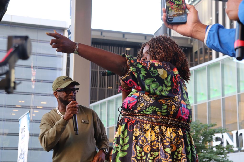 A person wearing a colorful, patterned dress gesturing while a man in a khaki hat holds a microphone in front of them.