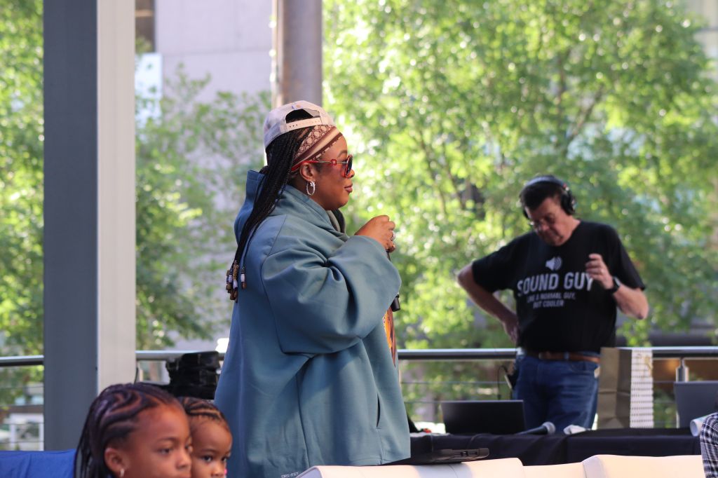 A person wearing a blue coat and headscarf standing in front of a person wearing a "Sound Guy" t-shirt, with two children seated in the foreground.