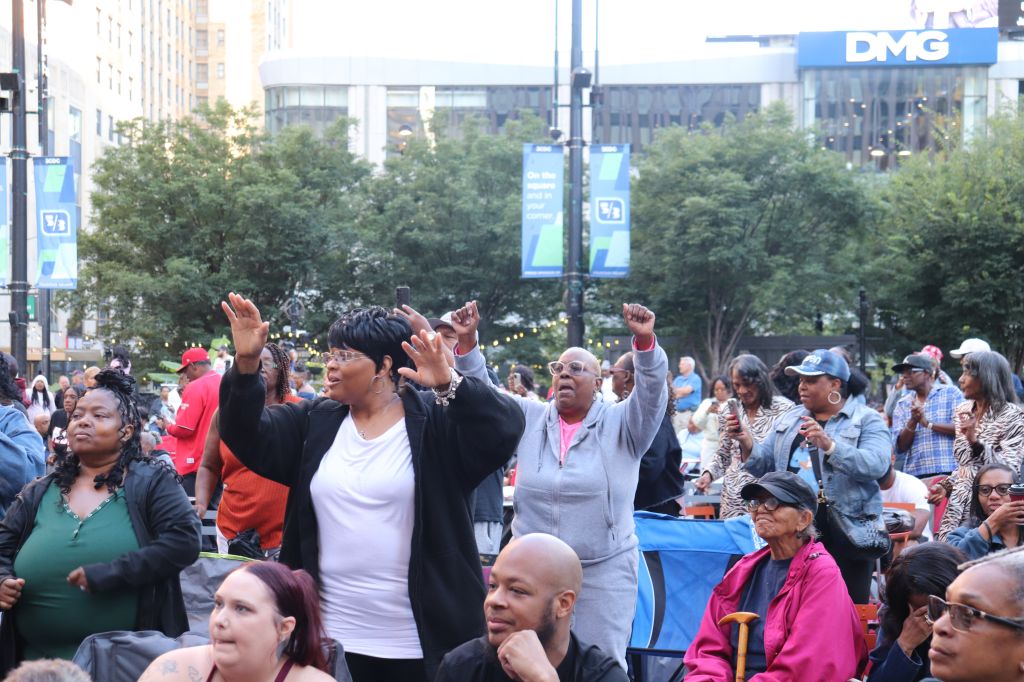 A crowd of people gathered in an urban setting, some raising their hands in protest or celebration. A DMG sign is visible in the background.