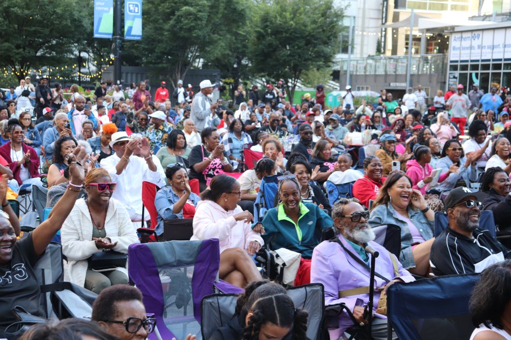 A large crowd of diverse people, many in wheelchairs, gathered in an outdoor public space, some waving and cheering.