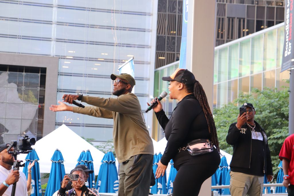 Two people speaking into microphones on a stage, with a backdrop of blue umbrellas and a building in the background.