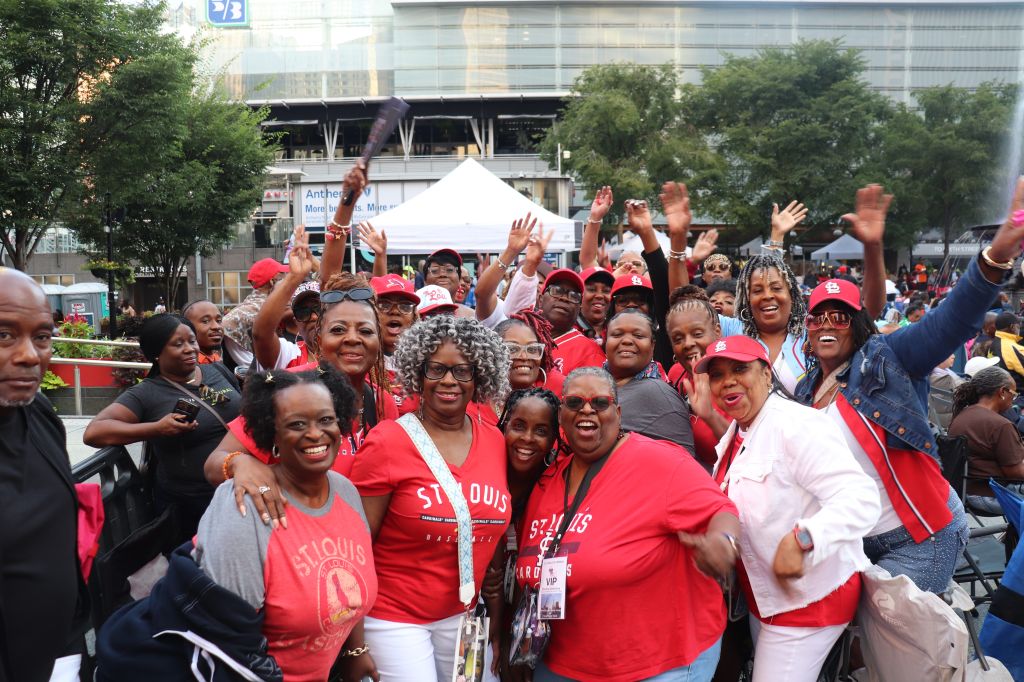 A large group of people wearing red St. Louis Cardinals baseball team shirts and hats, cheering and raising their hands in celebration.