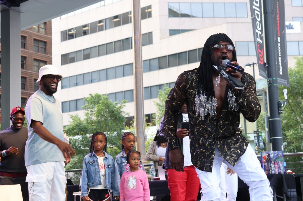 A group of people, including a man with dreadlocks speaking into a microphone, performing on an outdoor stage in front of a city skyline.