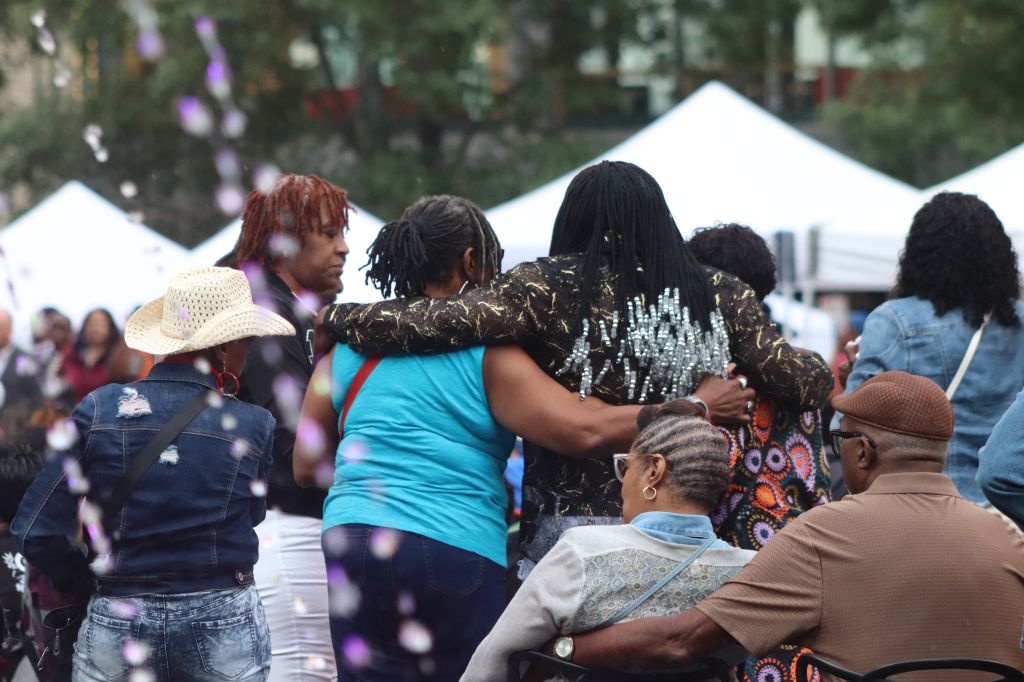 A group of people embracing and celebrating at an outdoor event, with tents and decorations visible in the background.
