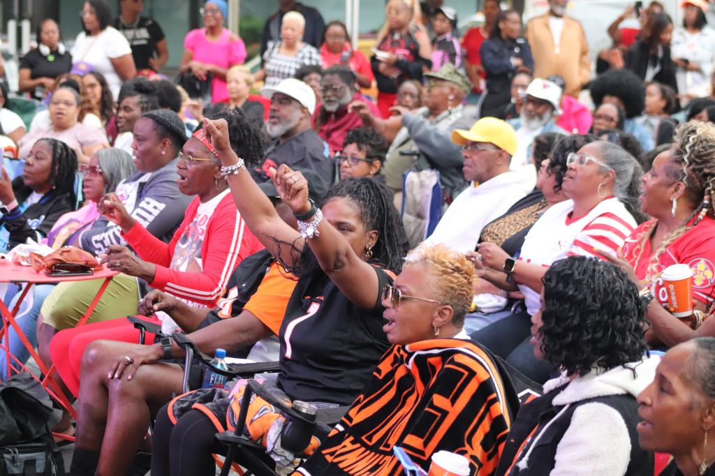 A large crowd of people, many wearing red and orange clothing, gathered together and cheering enthusiastically.