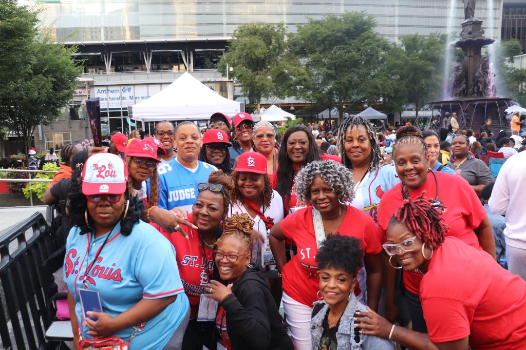 A large group of people wearing St. Louis Cardinals baseball team apparel and hats, gathered together in an outdoor setting with a fountain in the background.