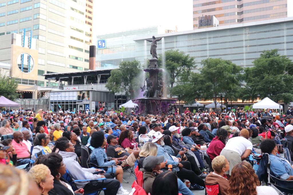 Crowded public plaza with a central fountain statue, surrounded by people seated on the ground watching a performance or event.