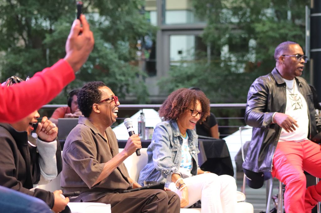 A group of people, including a man holding a microphone, laughing and interacting on an outdoor stage surrounded by trees.