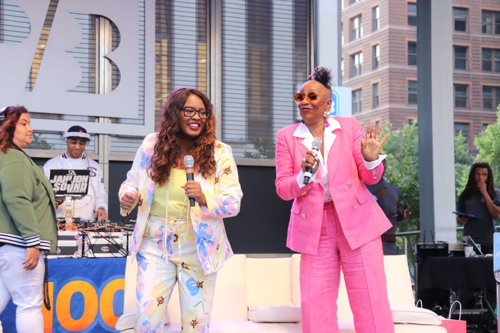 Two women on a stage, one wearing a colorful patterned outfit and the other a bright pink suit, holding microphones and speaking into them.