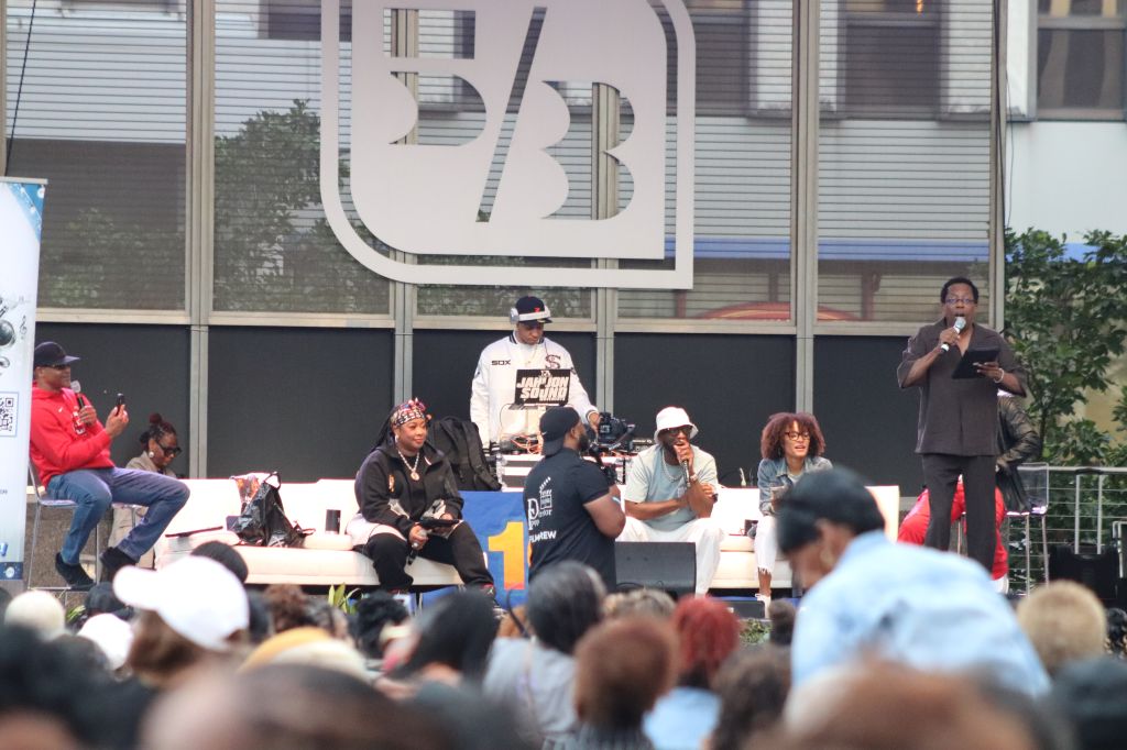 Outdoor event with a crowd gathered around a stage, where several people are speaking into microphones. The stage has the "BET" logo prominently displayed.