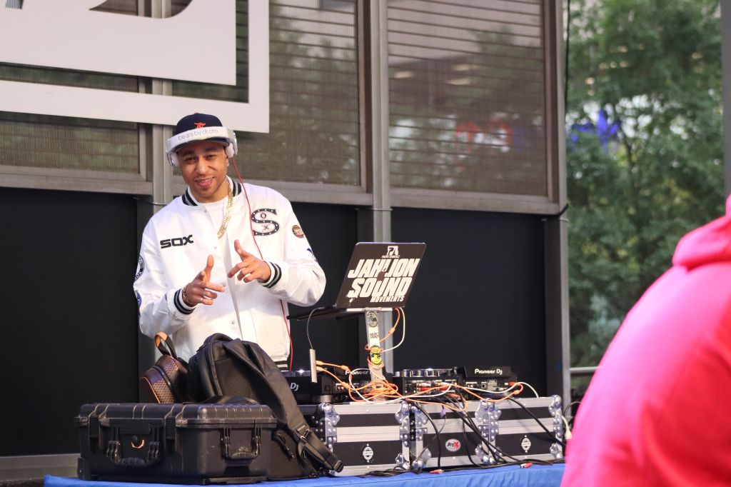A person wearing a white baseball jersey with "Sox" printed on it, holding DJ equipment and standing in front of a sound system.