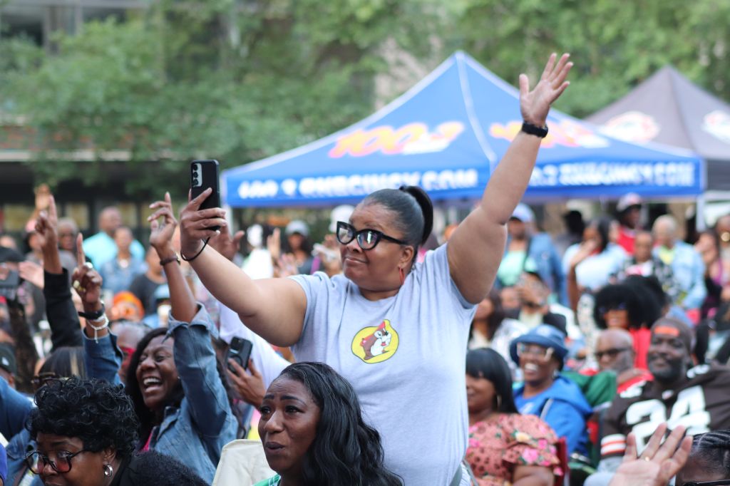 A crowd of people at an outdoor event, with a person in the foreground raising their arms and holding a smartphone to take a selfie.