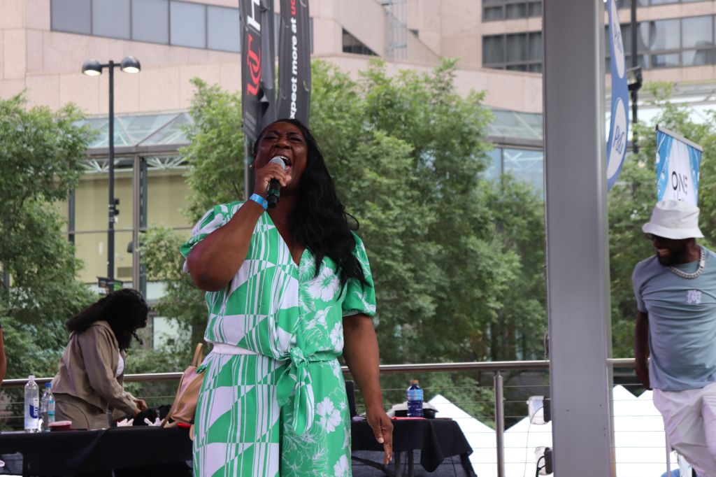 A woman with long dark hair singing into a microphone on a stage, surrounded by trees and buildings.