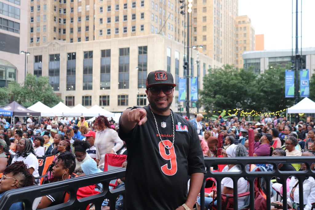 A smiling man wearing a Cincinnati Bengals jersey and cap stands in a crowded urban setting with tents and people in the background.