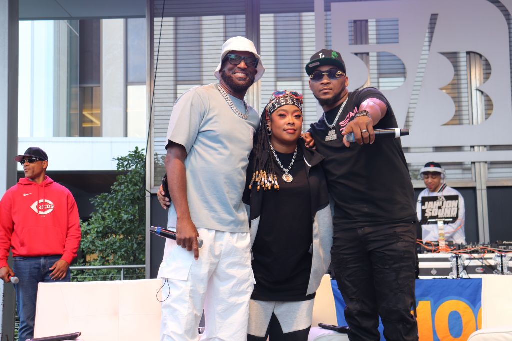 Three people posing together outdoors, wearing casual urban clothing and accessories. One person is wearing a white hat, another a black hat, and the third has a patterned headscarf. The background includes a building with signage.