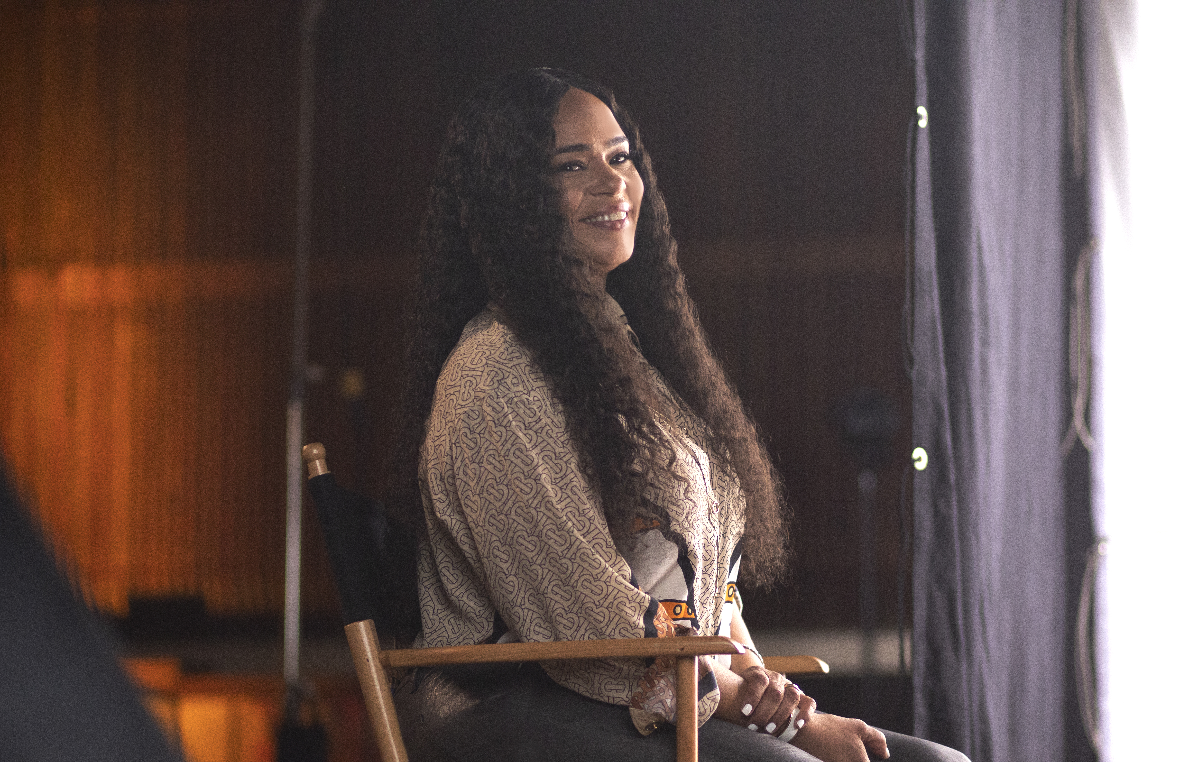 A smiling woman with curly dark hair wearing a patterned coat and sitting in a dimly lit room.